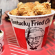 Bucket of KFC chicken, KFC mashed potatoes and gravy, and KFC cole slaw on red tabletop